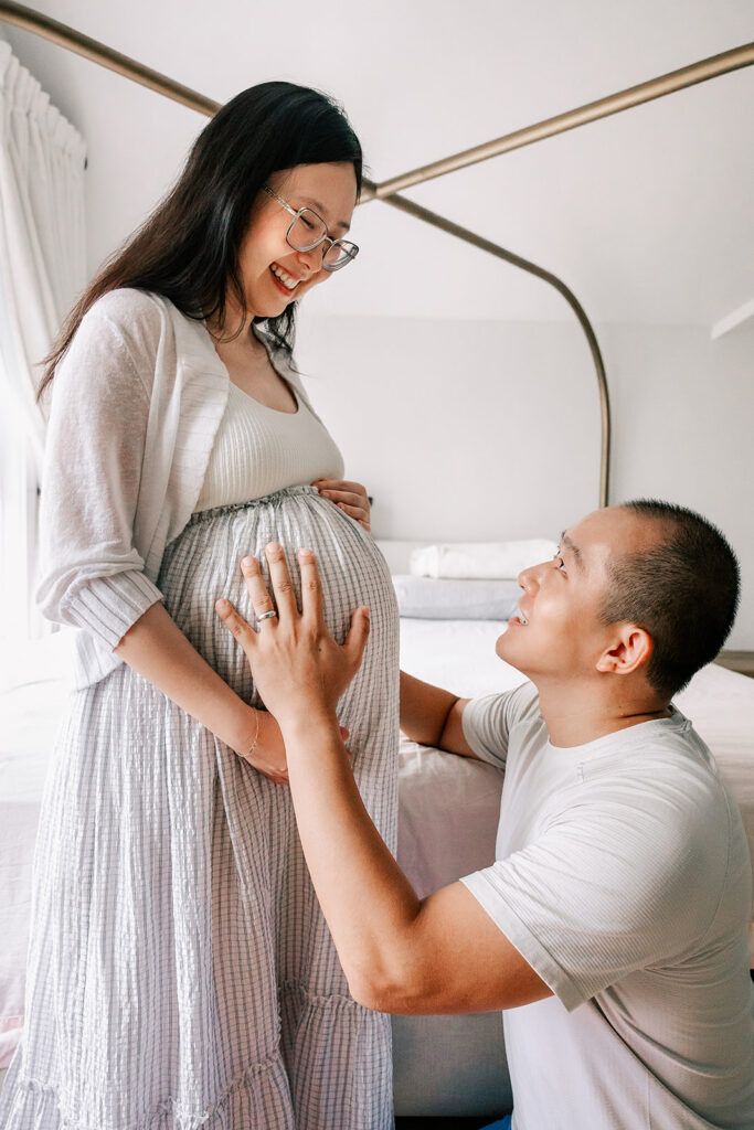 A soon to be mom smiles down on her bump while her husband kneels next to her with his hand on it after exploring baby shower venues in cambridge, MA.