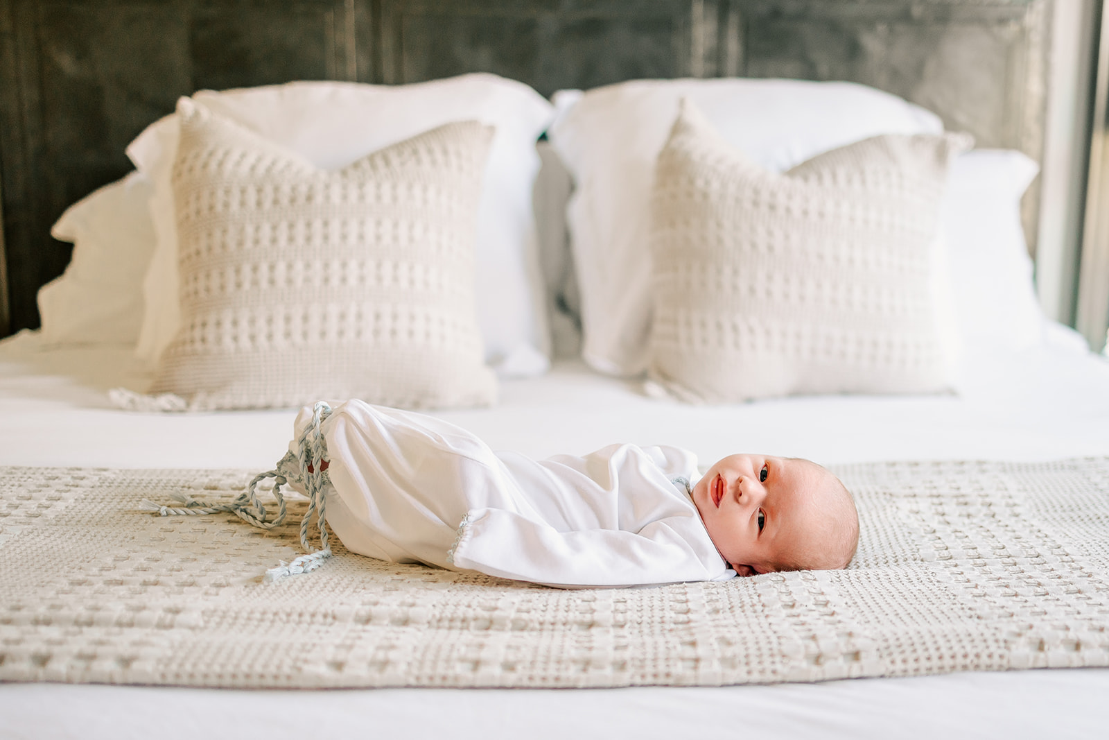 A newborn swaddled in a white blanket lays on a white bed while glancing toward the camera.