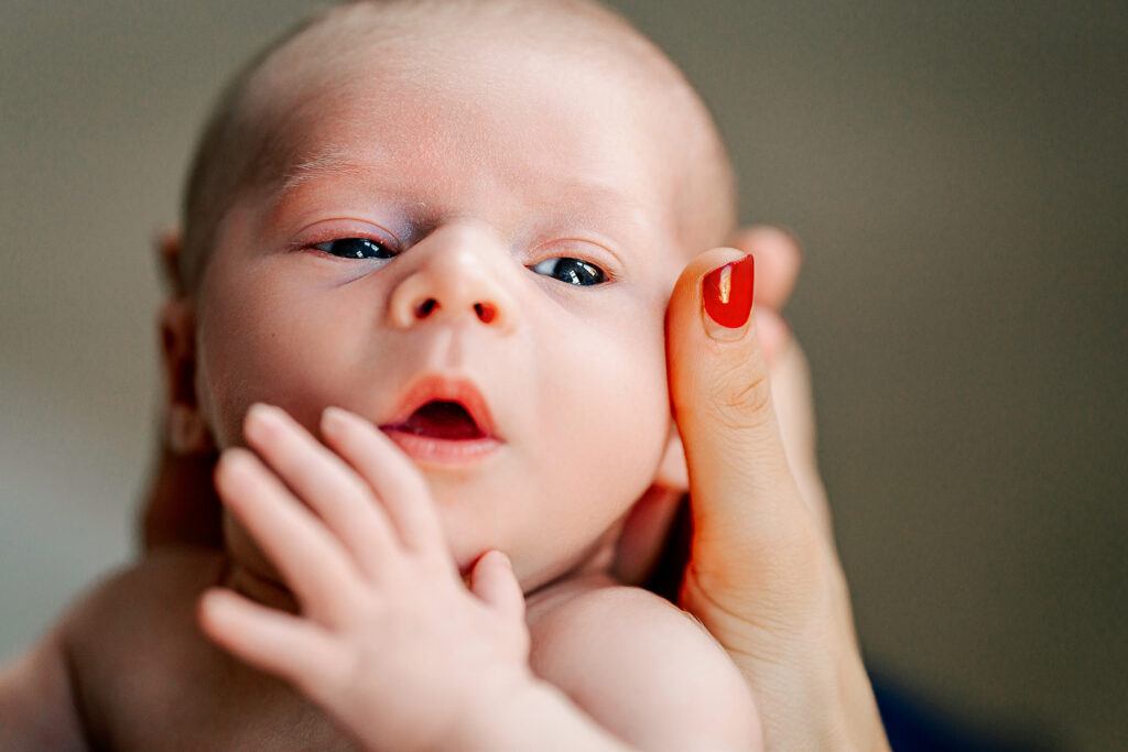 A close up shot of a newborn captures a mothers hand around the babies head with her thumb resting on the face of the baby before meeting the pediatric chiropractor in Boston.