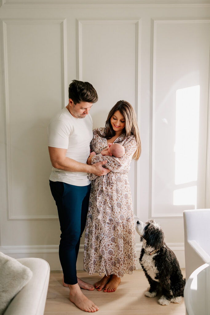 New parents stand holding their newborn in front of a white wall with their doodle sitting next to them after meeting a pediatric chiropractor in Boston.