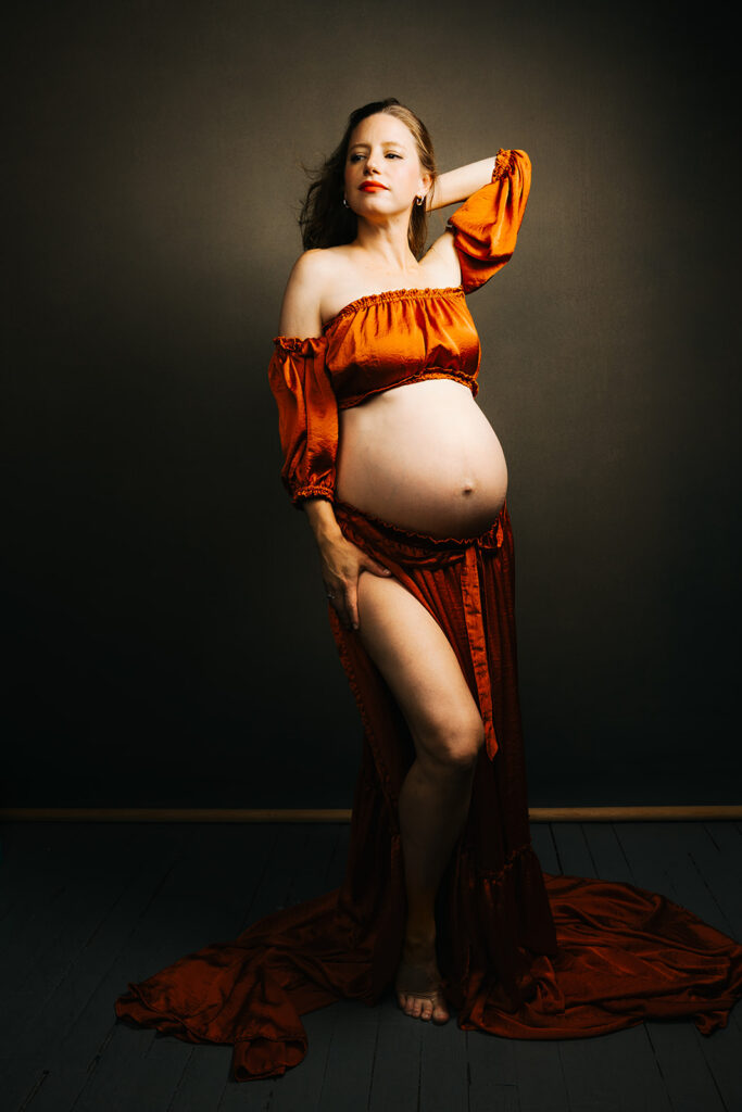 A pregnant lady stands with her leg out and a hand in her hair while looking into the distance of the studio.