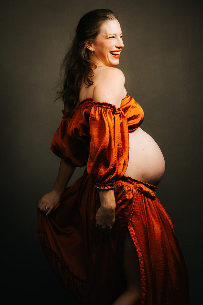 A laughing pregnant woman stares into the distance while standing in a orange two piece maternity outfit.