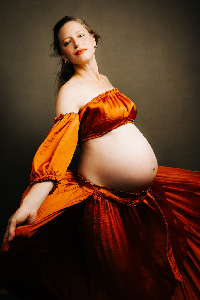 A pregnant woman twirls while playing with the train of her orange maternity skirt in a studio after enjoying pelvic floor therapy in Cambridge, MA