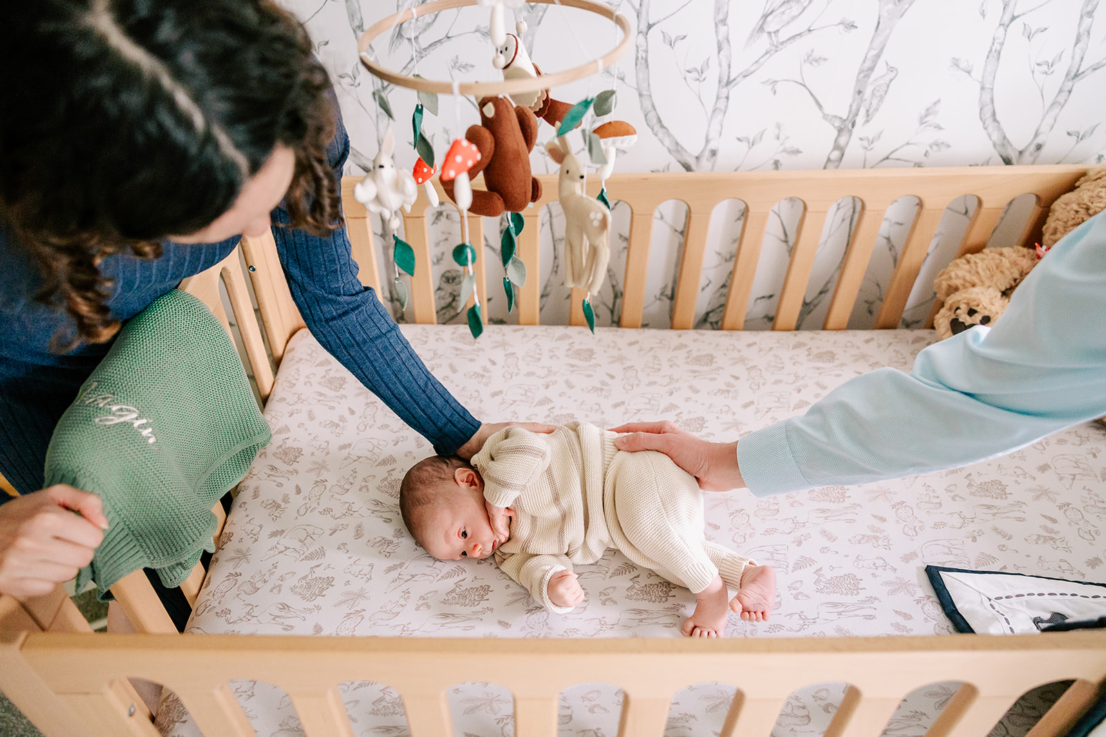 A newborn lays in a crib while mom and dad rest their hands on the baby.