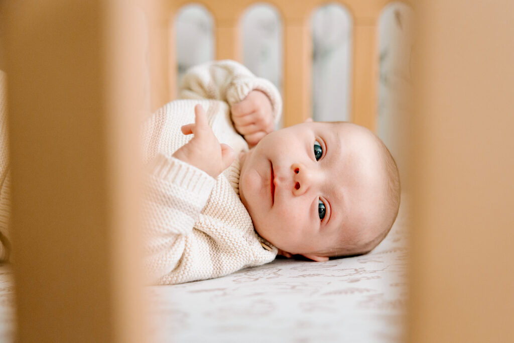 A close up of a laying newborn in it's crib in a tan onesie, eyes open.