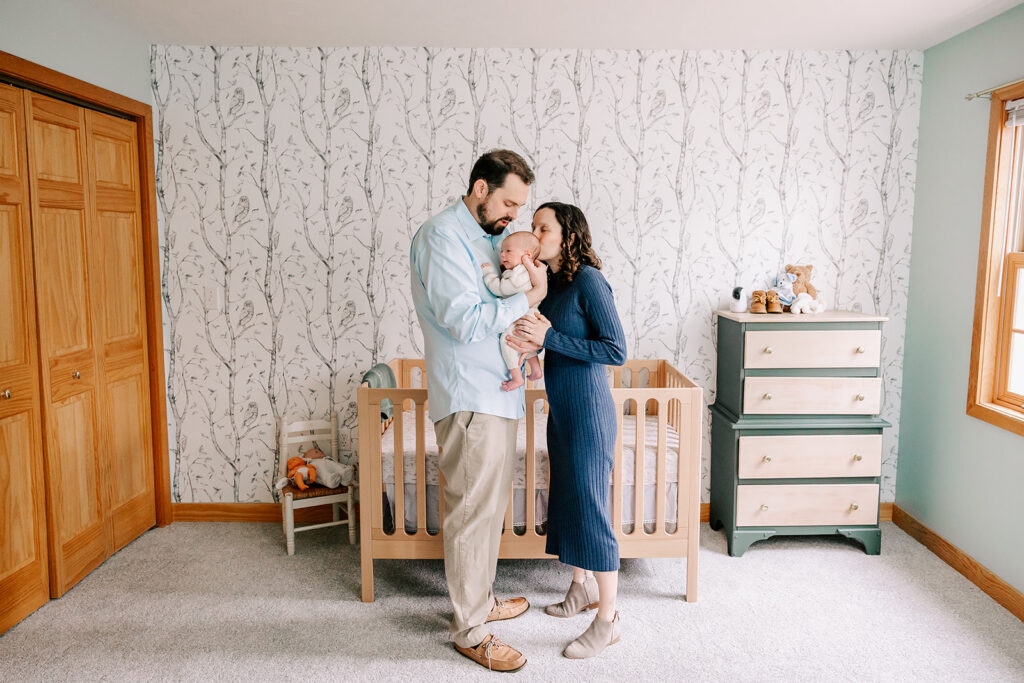 A new mother kisses the head of her newborn in dad's arms while standing in a nursery after meeting sleep consultants in Boston