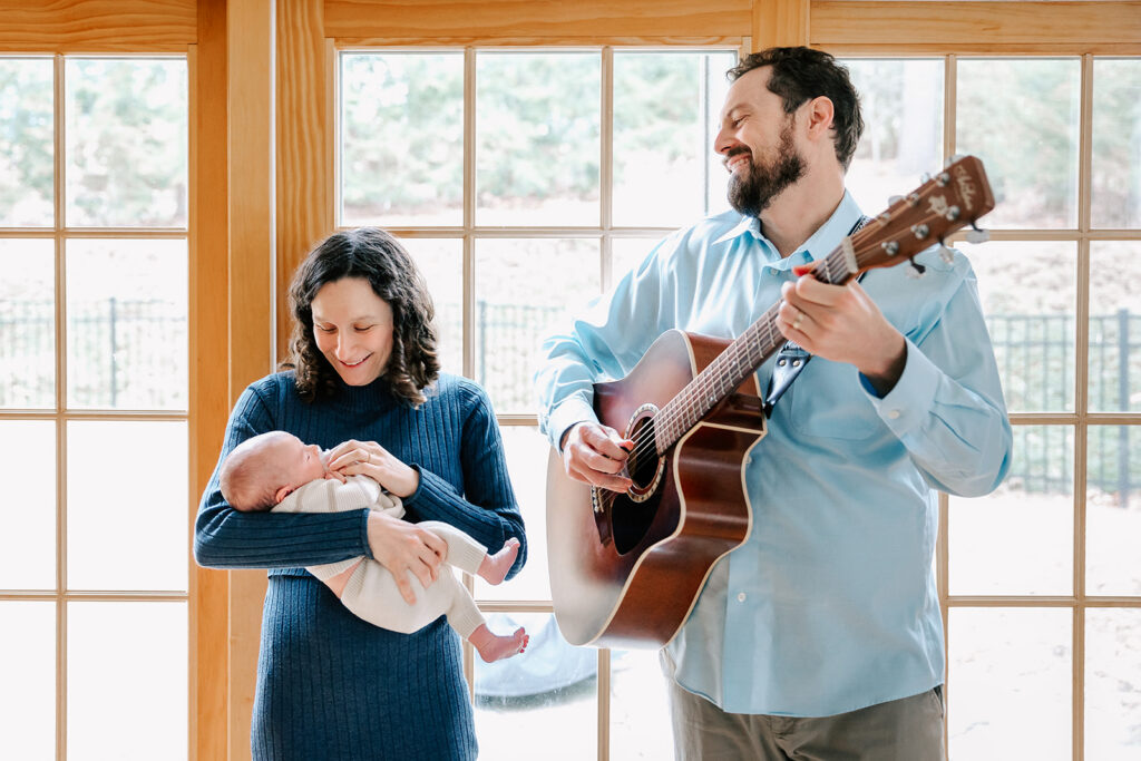 A mother holds a newborn in front of a window while her husband plays guitar next to them after meeting with a sleep consultant in Boston.