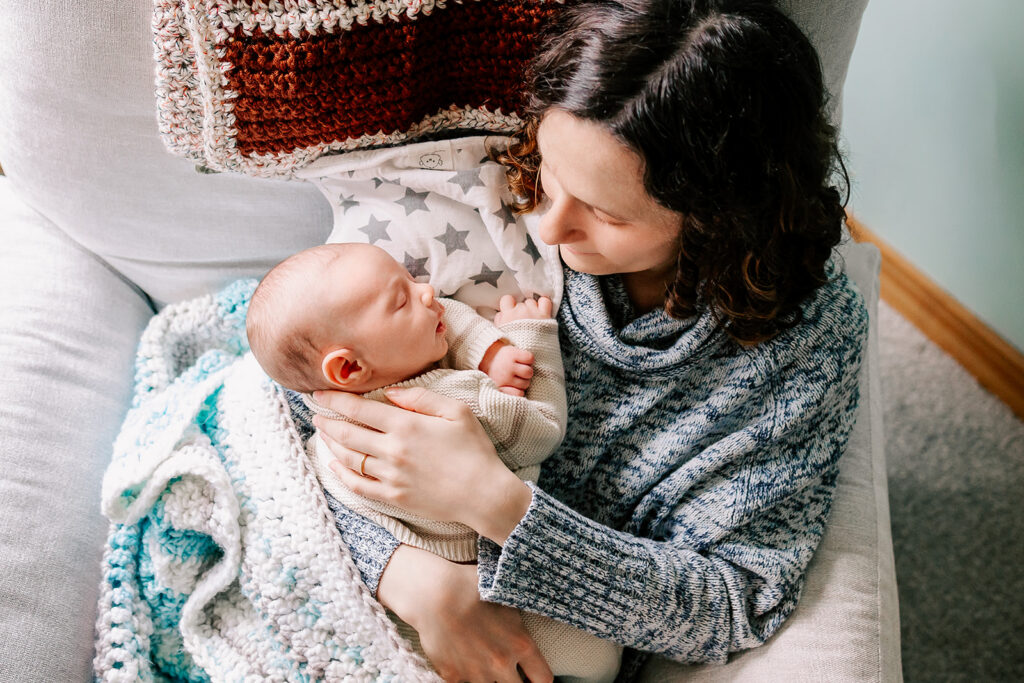 A mother looks down on her sleeping swaddled newborn after visiting a sleep consultant in Boston in a chair.