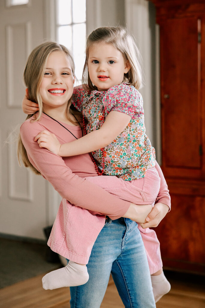 A young girl in a pink shirt smiles while holding her younger sister on her hip