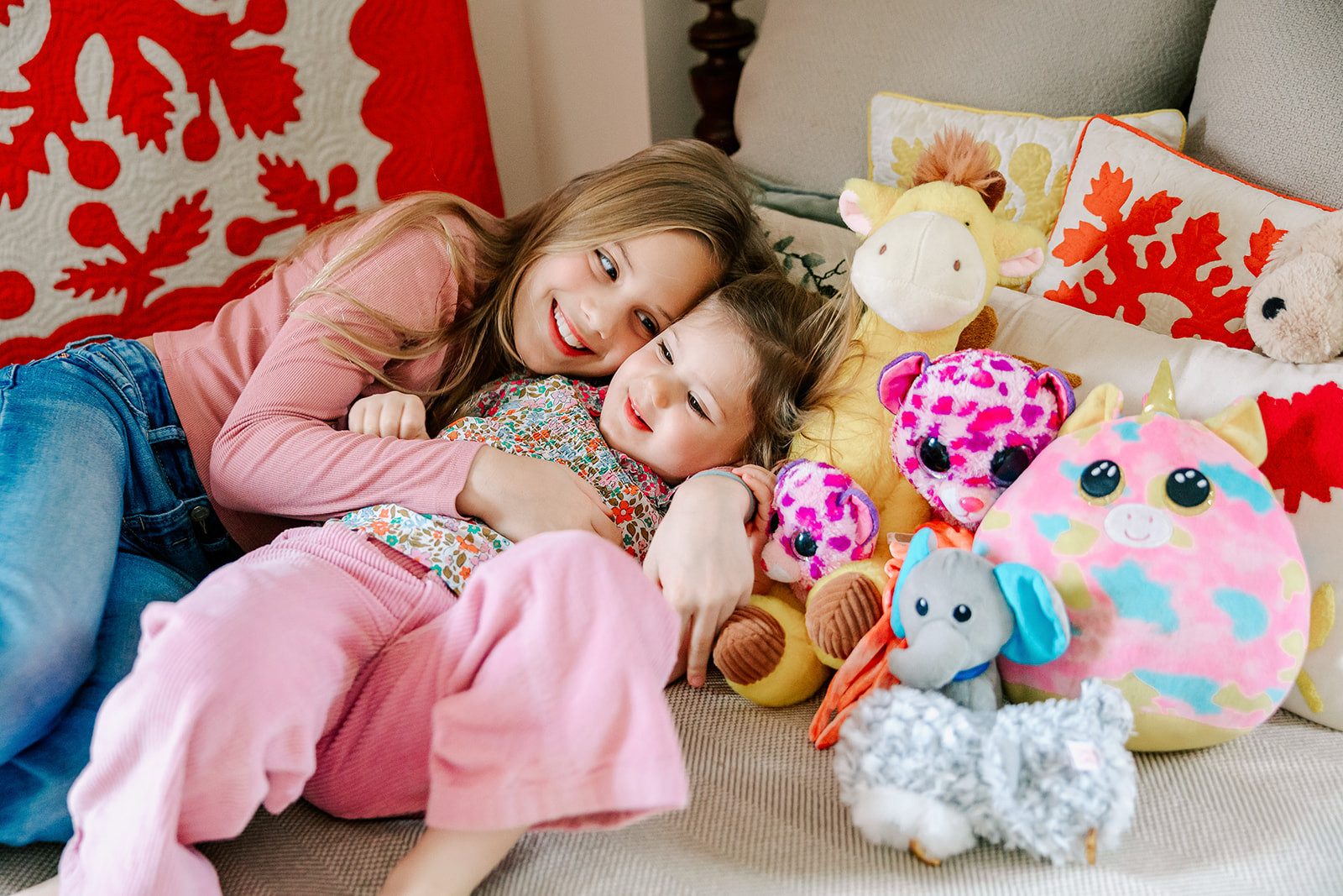 A young girl giggles and snuggles her toddler sister in a pile of stuffed animals in pink outfits