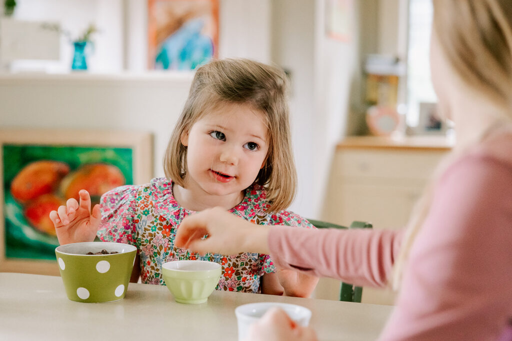 A toddler girl eating a snack in a bright floral print shirt at a kitchen table with mom's help after attending toddler yoga in Boston