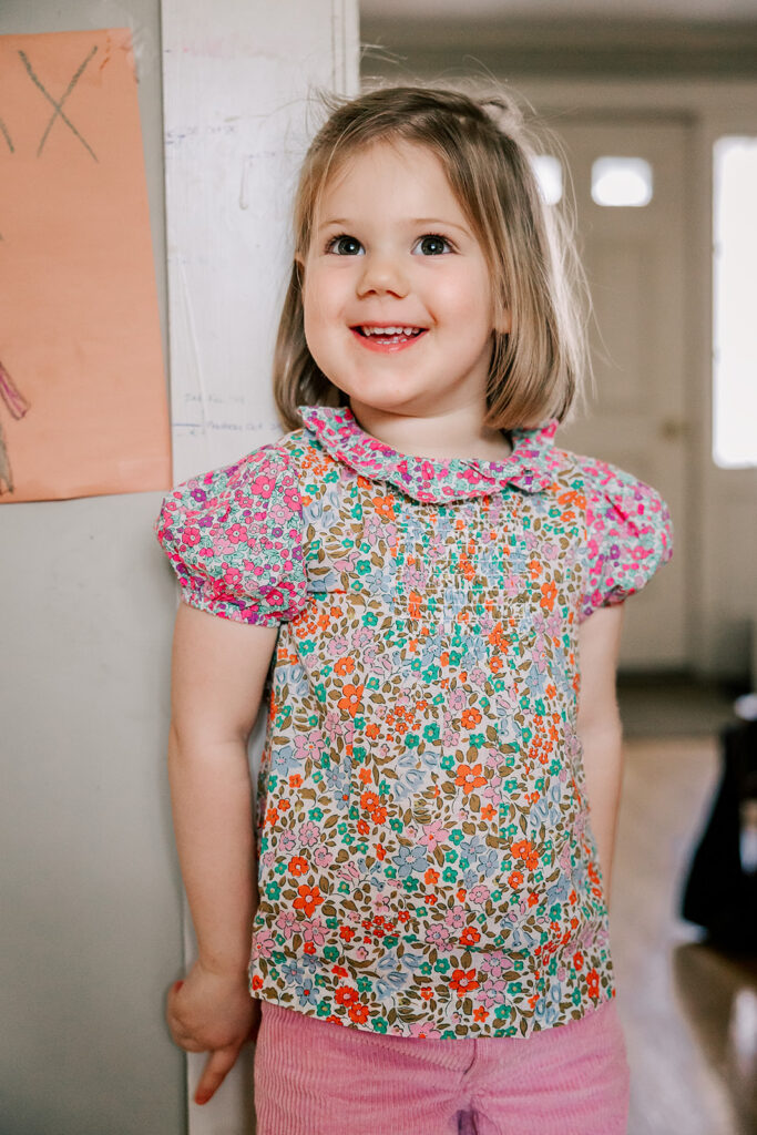 A toddler girl smiles while leaning on a kitchen wall in a bright flower print shirt after enjoying some toddler yoga in Boston