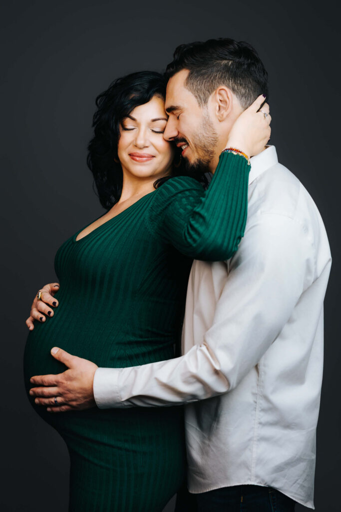 Mother-and-father-to-be snuggle against each other against a grey studio backdrop