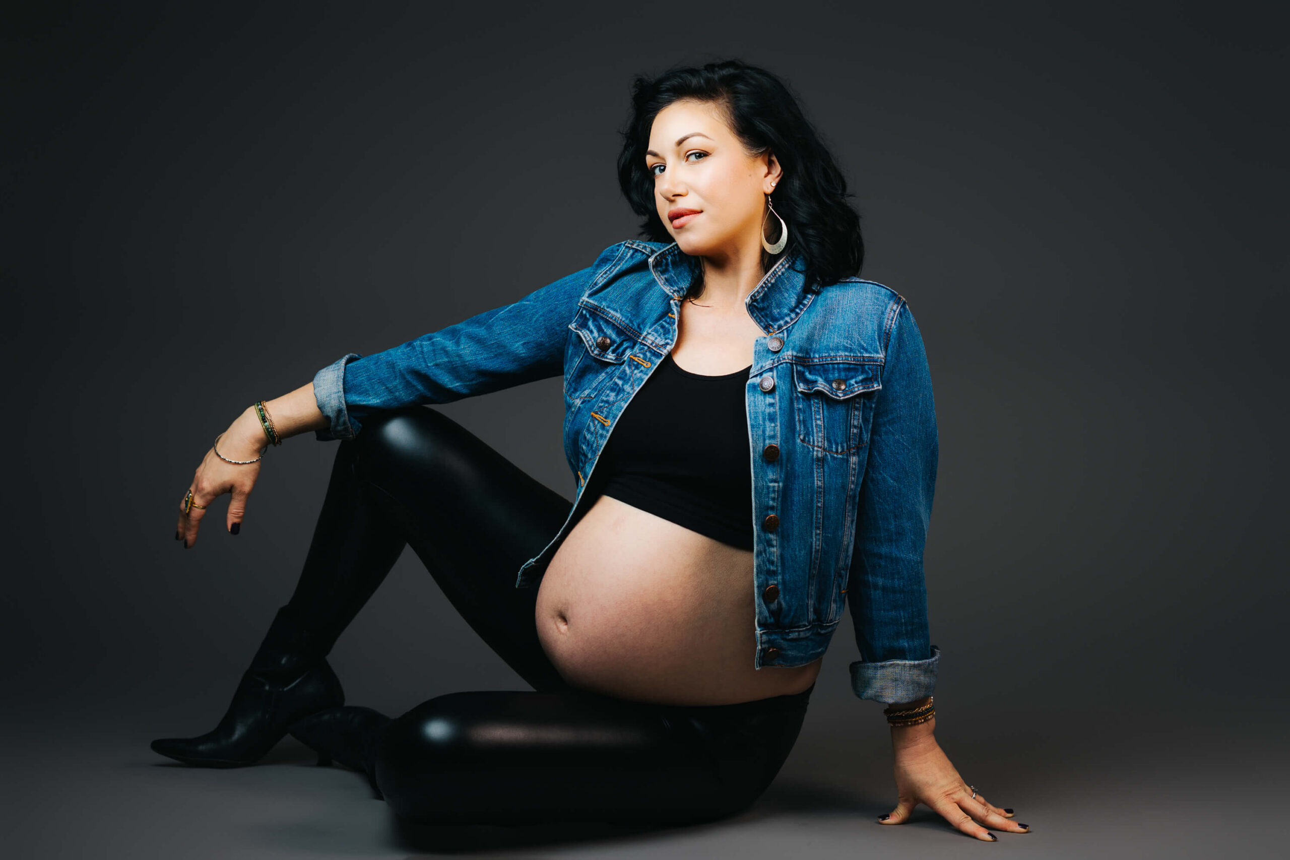 Mom-to-be lounges on the floor in black leather pants and a jean jacket