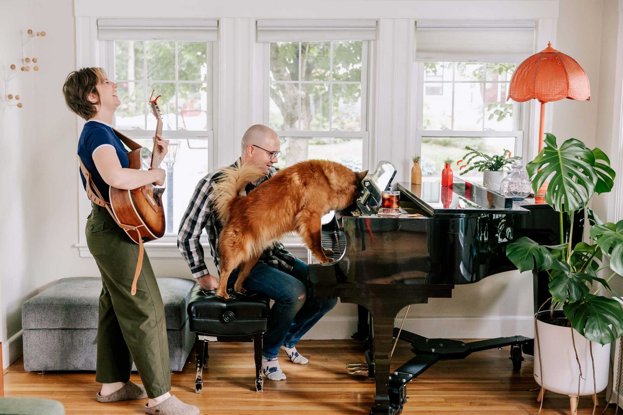 Man sits at a piano as a large brown dog jumps onto the seat and puts his paws on the keys; a woman standing next to the bench is playing the guitar and laughing