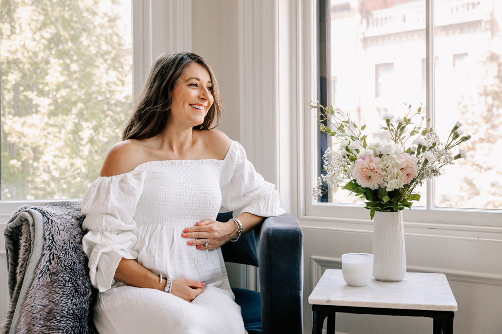 A smiling pregnant woman sits in a chair looking out the windows while holding her bump in a white dress after exploring Beehive Moms