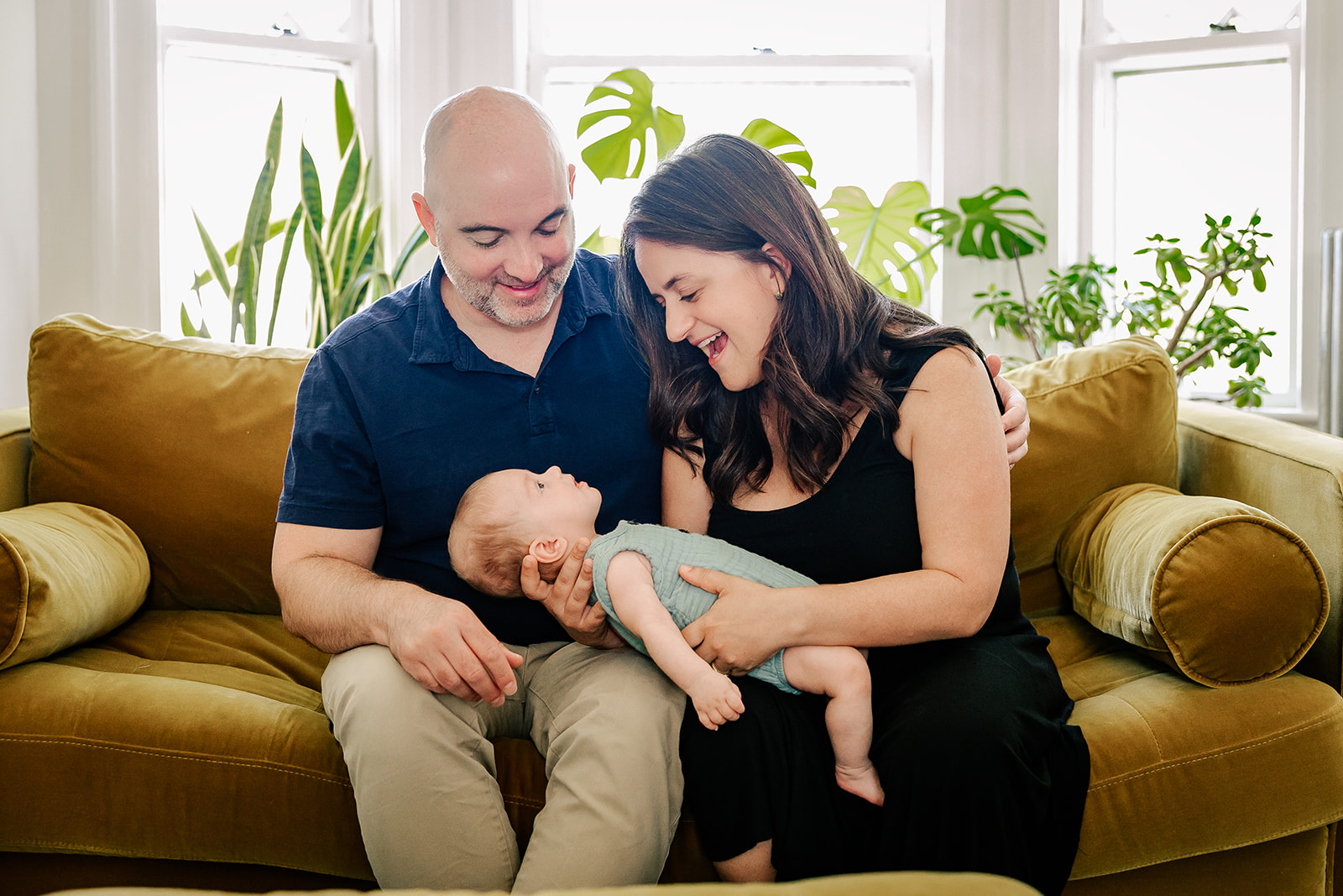 Happy mom and dad hold their happy baby boy in a green onesie in their laps while sitting on a couch after visiting Joy Street Boston