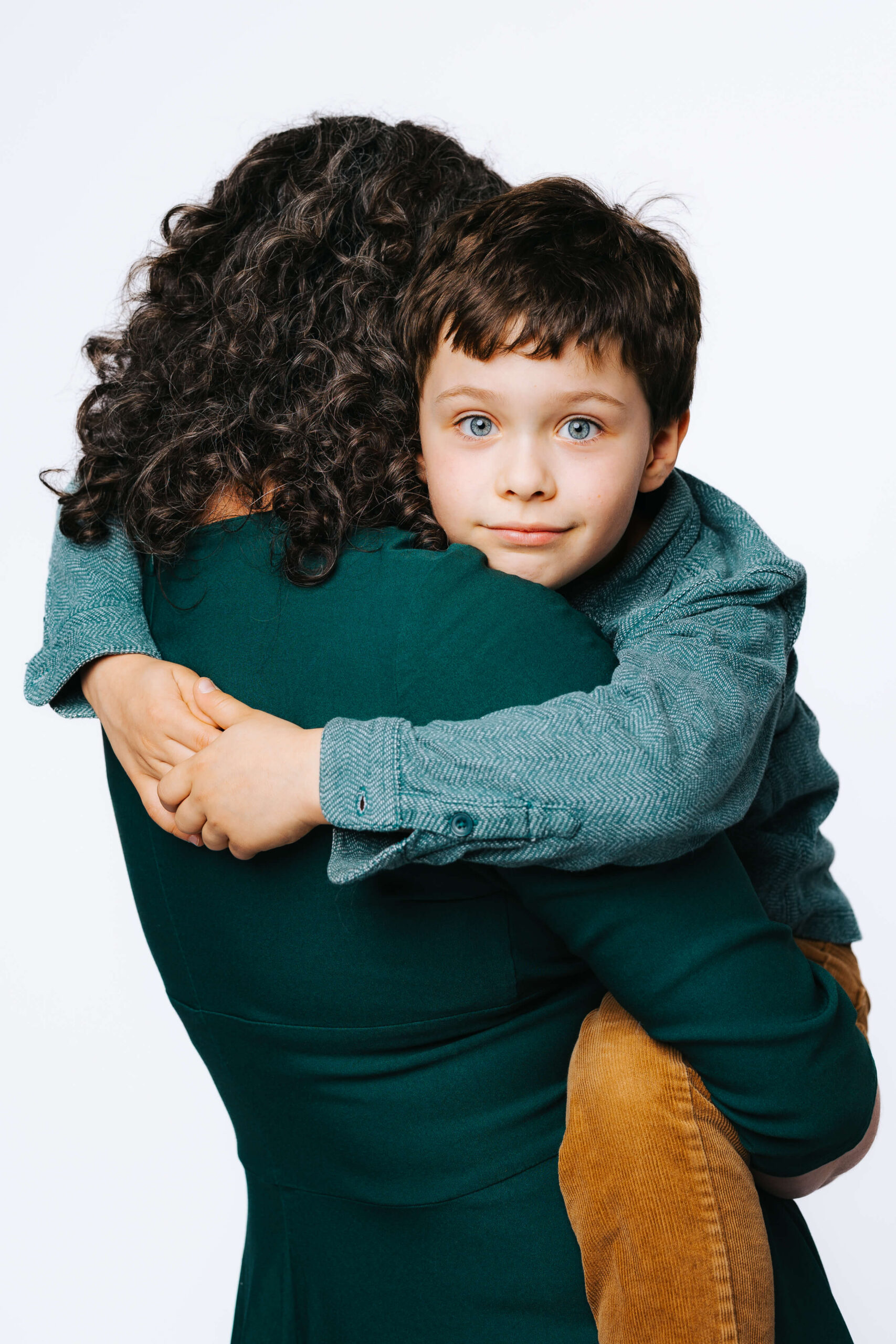 7 year old boy peeks over his mother's shoulder, his arms wrapped around her neck. He has a soft smile and bright blue eyes.