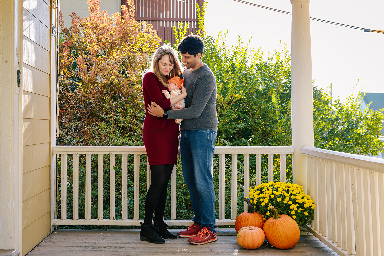 Happy new parents stand on their porch at sunset snuggling their newborn between them in red and grey